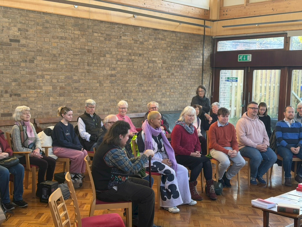 A quarter section of the meeting room with people posed as if in a meeting for worship, where people generally sit in silence waiting. People sit around the small altar like table in the middle, forming a circle with chairs spaced around it. Often there will be an empty chair on one or both sides of you, depending on how many people show up. A quarter section of the meeting room with people posed as if in a meeting for worship, where people generally sit in silence waiting. People sit around the small altar like table in the middle, forming a circle with chairs spaced around it. Often there will be an empty chair on one or both sides of you, depending on how many people show up.